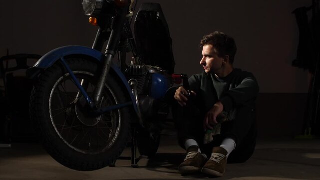 Owner taking a break from working on his old 1970s motorcycle, enjoying a cold beverage in a dark garage, embodying a classic biker lifestyle