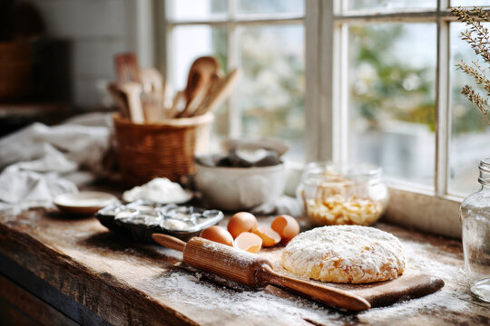 Bread dough resting on a flour dusted wooden board with rolling pin and eggs in a bright rustic kitchen window