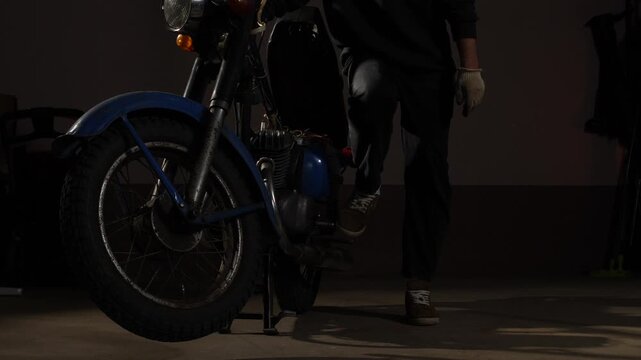 Mechanic wearing work gloves preparing to fix a classic blue motorcycle, highlighting the dedication to restoring vintage machinery in a dimly lit workshop environment