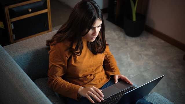 Young Hispanic woman sitting on her sofa looking angrily at her laptop screen, representing frustration, digital problems, work challenges and emotional reactions during technology use at home