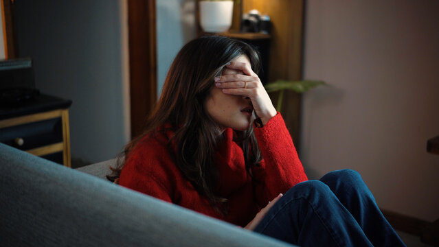 Young Latina woman covering her face in frustration while sitting in the living room of her home, representing stress, disappointment, emotional exhaustion and personal challenges in everyday life