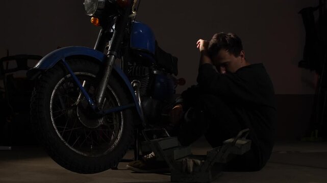 Young man feeling frustrated and stressed, sitting on the floor leaning against a broken vintage blue motorcycle, a toolbox lying open nearby in a dimly lit garage