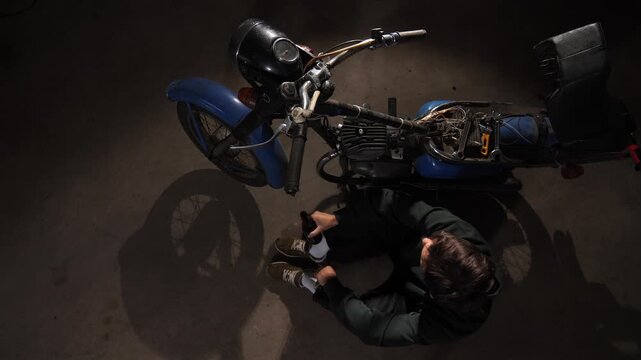 Man pausing from repairing a vintage motorcycle in a dark garage, sitting on the floor and drinking from a bottle, representing work life balance and refreshment after focused labor