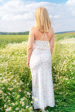 Blonde woman, with her back to camera, wearing long white sundress with floral print, stands in middle of chamomile field against backdrop of green meadow and cloudy sky, feeling free and calm