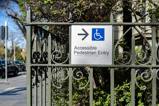 Accessible pedestrian entry sign with wheelchair symbol on wrought iron fence of a historic Victorian-era building in Melbourne, Australia, heritage architecture adapted for modern disability access