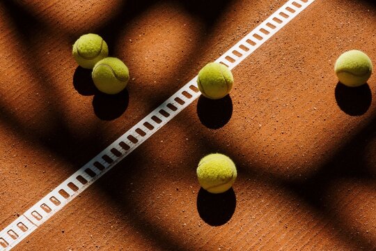 Tennis balls rest on a clay court surface marked by sharp shadows cast from a nearby fence.