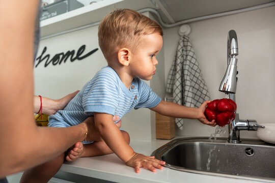 Mother helping toddler son wash bell pepper at sink