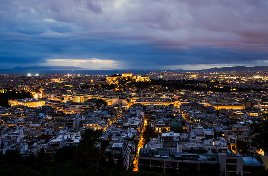 Panoramic night view of Athens city, Greece with the illuminated Acropolis and Parthenon visible on the hill. City lights, the sea in the background, and the iconic ancient monument