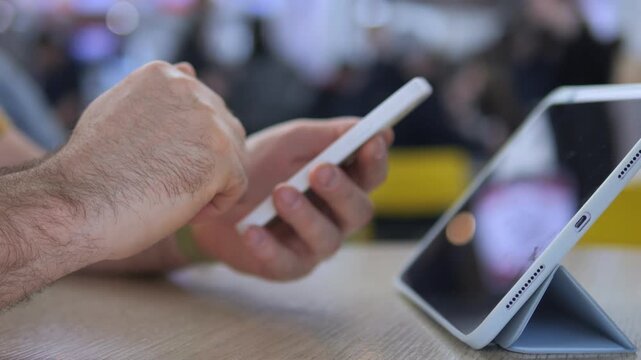 Close-up of hands using smartphone and tablet. Focus on fingers scrolling and tapping. Blurred background shows public setting. Suitable for tech, communication, or multitasking themes