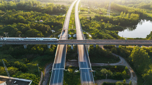An aerial view of a high-speed train traveling on an overpass crossing a highway through lush green landscape with water bodies.