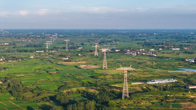 High voltage transmission pylons stretch across a lush rural agricultural landscape with scattered settlements under a clear sky.