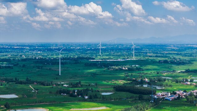 Wind turbines are set in a sprawling green rural landscape with fields, ponds and scattered settlements under a cloudy blue sky.