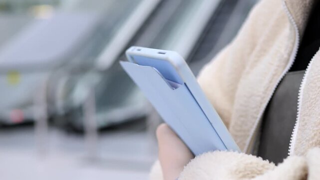 Close-up of hand holding blue smartphone. Person wears light-colored fleece jacket. Background shows blurred escalator and indoor space. Focus on device and casual travel setting