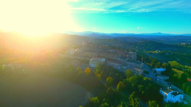 Stupendous aerial push-in shot from Monte Rinaldo, approaching the town centre with its vintage buildings while the sun shines from the left, piercing through mountains, with azure sky, hazy clouds