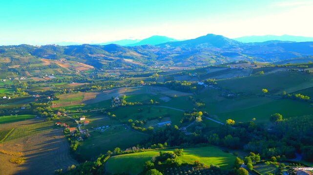 Refreshing aerial pullback pan right shot from Monte Rinaldo, from the Sibillini Mountains' majestic outlines in the distance to the hills, fields, meadows, roads, trees typical of Marche landscapes