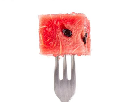 Fresh red watermelon cube with black seeds and water droplets pierced by a stainless steel fork, centered on a clean white background for food, nutrition, and summer concepts.