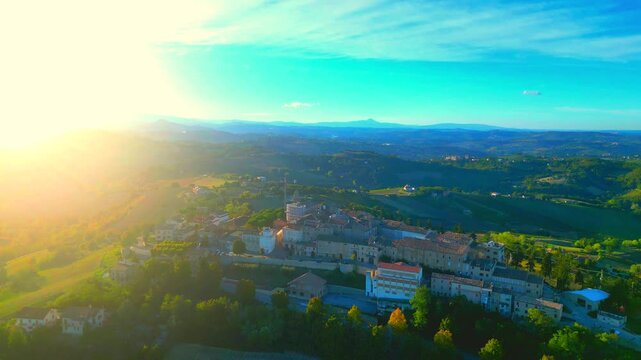 Pleasant aerial push-in tilt down shot from Monte Rinaldo, flying over the town centre and its vintage buildings while afternoon sunlight fills the left side of the picture, amongst Marche hillscapes