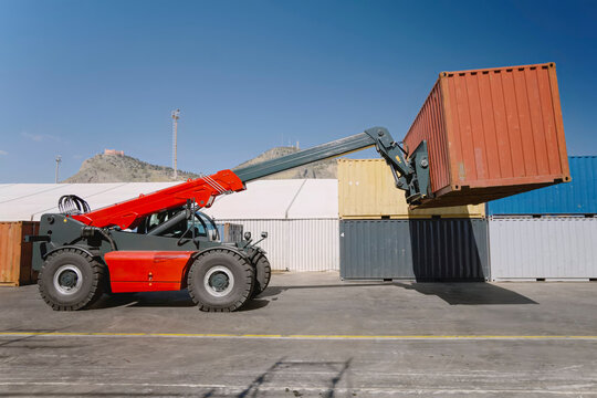 Red telehandler lifting an orange shipping container among stacked containers at a port yard