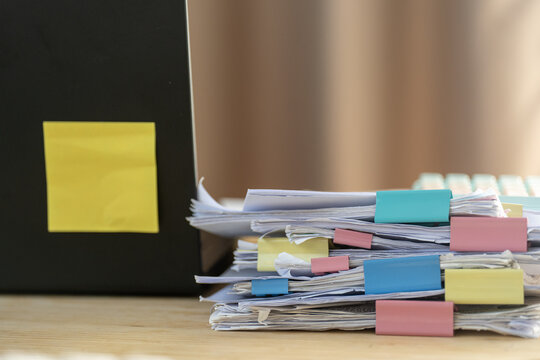 Stacked papers binded with colorful clips. Yellow sticky note sticks to dark folder beside. Background blurred, focus sharp on cluttered documents. Office scene suggests busy workload or pending tasks