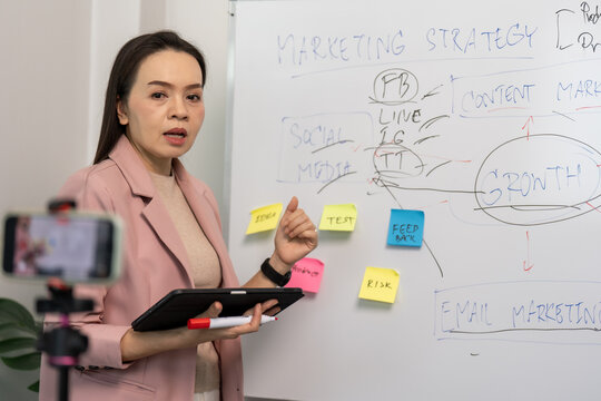 Woman teaches marketing strategies with a tablet and sticky notes. She points at a whiteboard with hand-drawn diagrams. Her expression shows focus and engagement