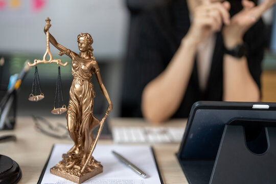 Golden statue of Lady Justice stands tall. She holds scales, symbolizing fairness and balance. Behind her, a person adjusts glasses, focused on work. Desk holds tablet, pen, and papers