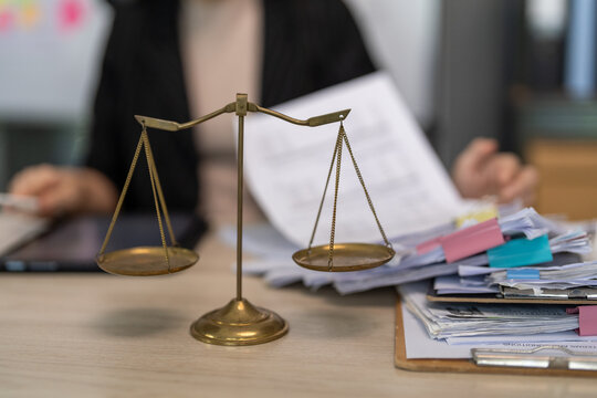 Golden scales sit on a cluttered desk. Hands hold papers, suggesting legal work or dispute resolution. Documents and colorful sticky notes pile beside the scales