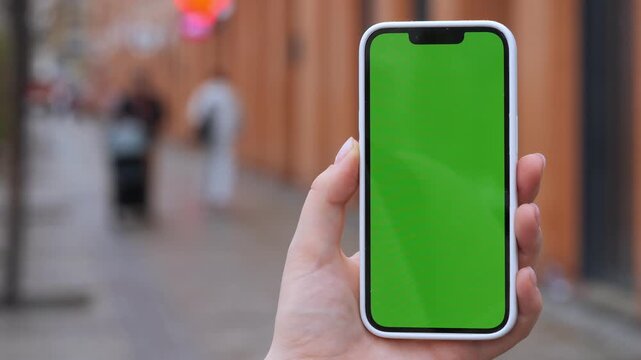 Hand holds smartphone with green screen. Background shows blurred urban street. Person appears neutral, focused on phone. Ideal for app mockups, digital marketing, or UI design projects