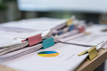 Fototapeta premium Pile of papers rests on wooden desk surface. Colorful clip holds documents neatly together. Pie chart sits among papers, hinting at data analysis