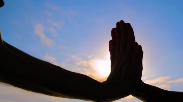 Close up shot: silhouettes of woman and man are giving a high fives hand gesture against the sunset or sunrise sky with sun lens flare. Friendship, celebration, cheerful and positive concept