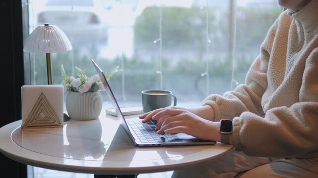 Person in cozy sweater typing on laptop at cafe table. Hands move steadily, focused on work or messaging. Soft light filters through window, creating calm ambiance
