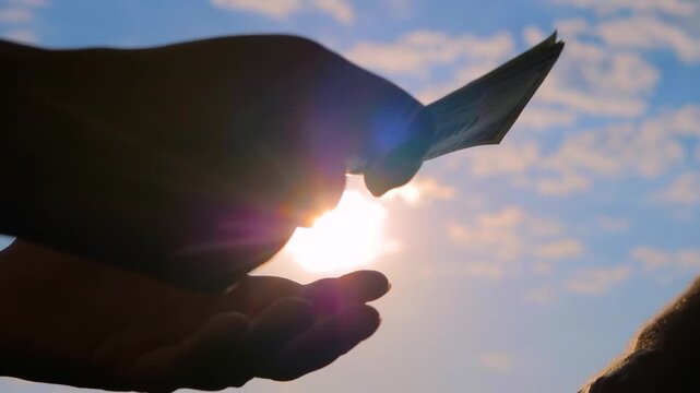 One person is giving a bribe to another person. Woman is counting dollar bills. Hands silhouettes with money, banknotes against the sunset, sunrise sky with sun lens flares, close up view
