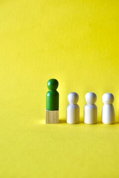 Leadership and career advancement concept with a green wooden pawn standing on a block at the head of a line of white figures on a yellow background.
