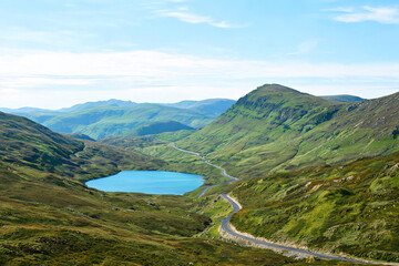 Fototapeta premium A scenic highland landscape featuring a turquoise lake, winding mountain road, and rolling green hills under a clear blue sky.