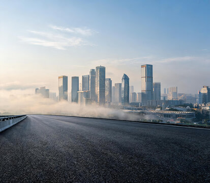 An empty asphalt road stretches forward, leading to a modern city skyline partially shrouded in mist under a clear blue sky.
