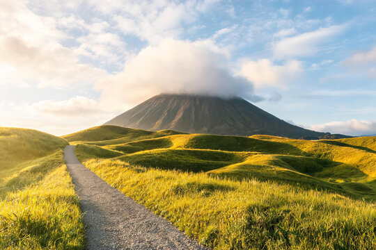 A volcanic peak partially shrouded in cloud rises behind rolling grassy hills with a narrow gravel path under a bright sky.