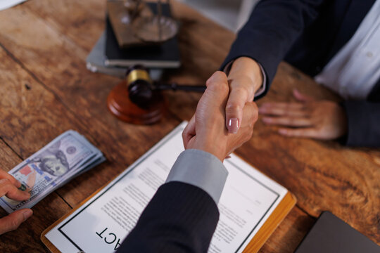 People shaking hands. Finalizing an agreement next to a contract. A stack of dollars. And a judge's gavel on a wooden table. Depicting themes of legal deals. Financial transactions