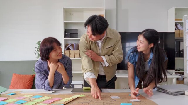 Three colleagues lean over a desk examining a hand-drawn business strategy map covered in colorful sticky notes, brainstorming project ideas together beside a laptop in a bright modern office.