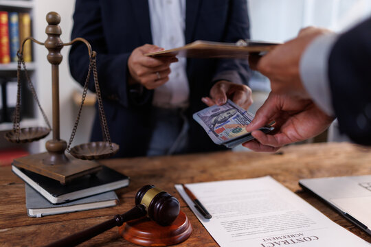 Hands engaging in a clandestine transaction, with a bundle of cash being passed to a legal professional holding a clipboard, placed on a desk with scales of justice, a gavel, and a contract