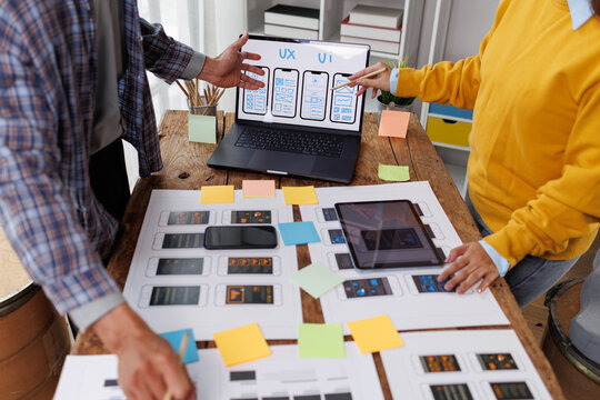 Digital product designers collaborating on ux and ui wireframes, developing mobile application interfaces on a laptop, tablet, and printed mockups, optimizing user experience and usability