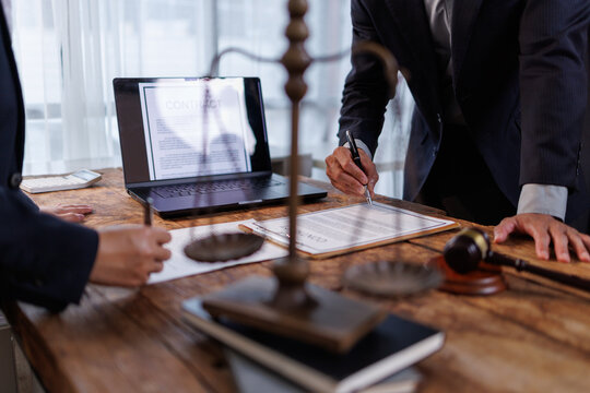 Legal professionals working together, reviewing a contract on a laptop and signing paper documents, with a gavel and scales of justice prominent on the office desk