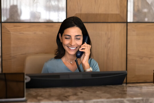 Smiling female receptionist working at hotel reception desk, talking on landline phone, handling guest service, make room reservations, taking booking, managing client inquiries. Hospitality services