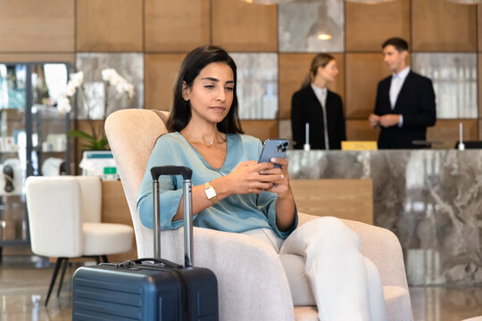 Woman checking smartphone sitting in hotel lobby with suitcase, using travel mobile application, processing check-in, reviewing booking reservation details. Business trip, digital services concept