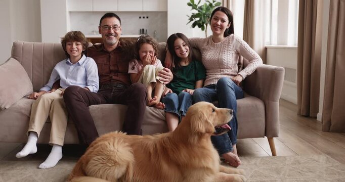 Cheerful Caucasian married couple sitting on couch with siblings, two daughters and son, posing for family portrait in living room at home, golden retriever dog lying on floor. Family bonding, love