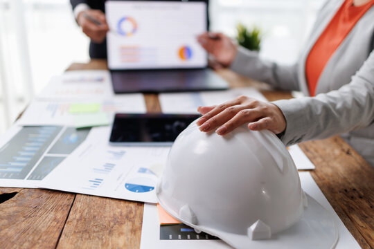 Business professionals collaborating at a wooden conference table. Reviewing financial data charts on a laptop while a safety hard hat rests on paper documents