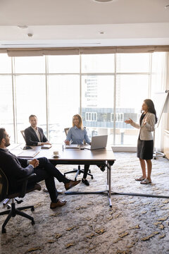 Multiethnic corporate team participate in group meeting or training led by female leader or coach stands in front of colleagues, explaining strategy, discussing ideas, business solutions. Presentation