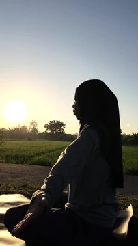 Slow motion vertical clip of a silhouetted hijabi woman doing a seated upward yoga stretch on a mat by a green rice paddy. Beautiful golden morning sunlight. Perfect for wellness themes.
