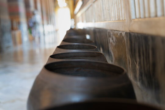 Row of bronze alms bowls along a wall inside Wat Pho temple in Bangkok, Thailand, used for Buddhist coin donation rituals.