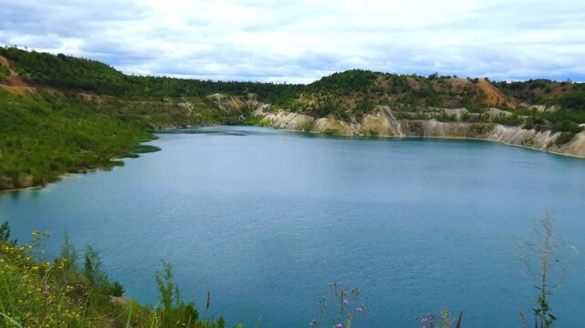 People walk along the edge of a large blue lake. The lake is surrounded by green trees and hills. The sky is overcast with some clouds above. Chalk quarries in the city of Volkovysk in Belarus, the Be