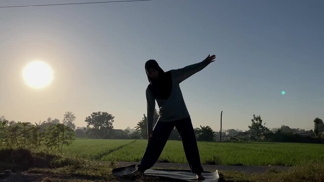 Slow motion silhouette of a Muslim woman in a hijab doing the triangle yoga pose (Trikonasana) on a mat outdoors. Beautiful golden sunrise over a lush green rice field. Perfect for wellness themes.
