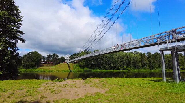 People enjoy walking on a bridge that spans over a river. The area is filled with trees and grass. The scene shows a bright day with clouds in the sky.  The suspension bridge in Mosty, Belarus, 